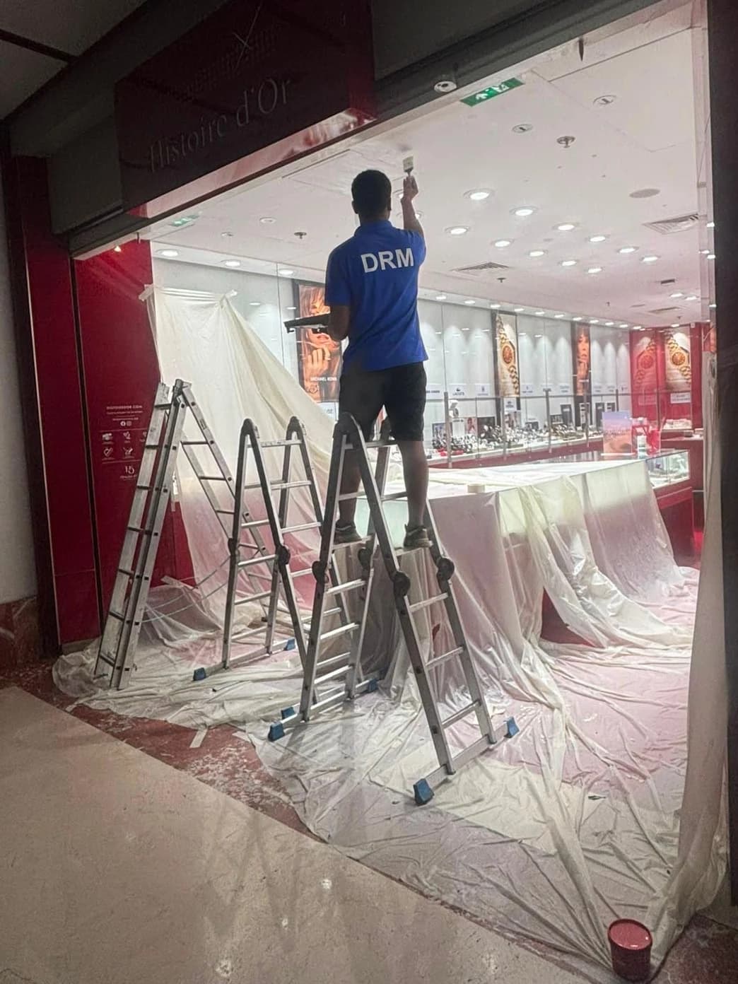 Technicien DRM sur escabeau à l’entrée d’une bijouterie bâchée, pendant la pose du rideau métallique